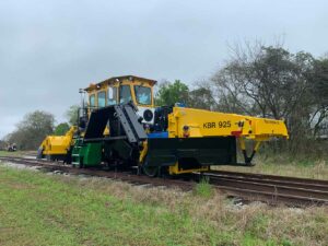 KBR 925 Ballast Regulator with Plow attachment on railway with green grass in front and green trees behind.