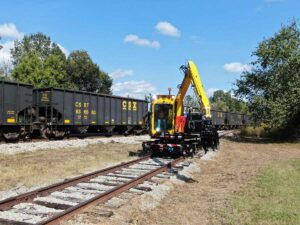 The KTC 1250 Tie Crane on railway tracks with railroad cars on another track behind it.