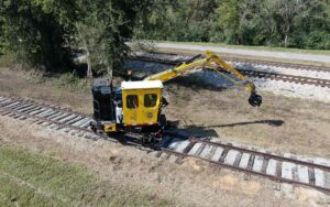 Piece of yellow railway machinery with attachment arm on railroad tracks with green trees in background.